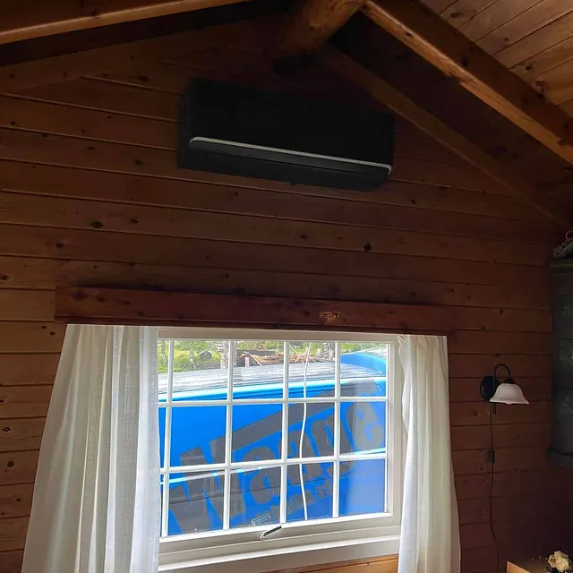 Sort varmepumpe montert høyt på veggen i hytte med panel og synlige takbjelker.