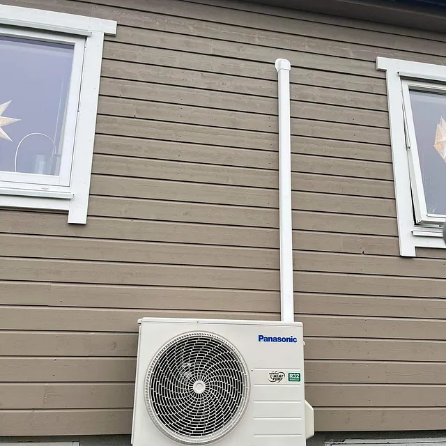 Panasonic utedel montert på brun trevegg mellom to vinduer med hvite karmer.