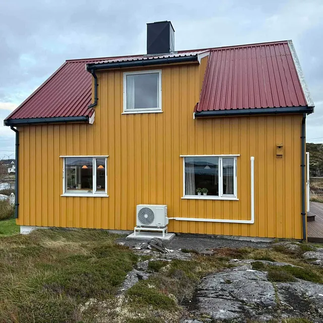 Gul enebolig med rød takstein og utedel av varmepumpe montert på grunnmur.