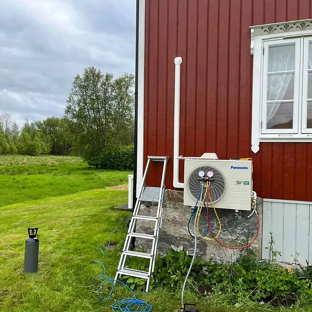 Panasonic utedel under installasjon på rød trevegg med hvite vinduskarmer og grønn hage.
